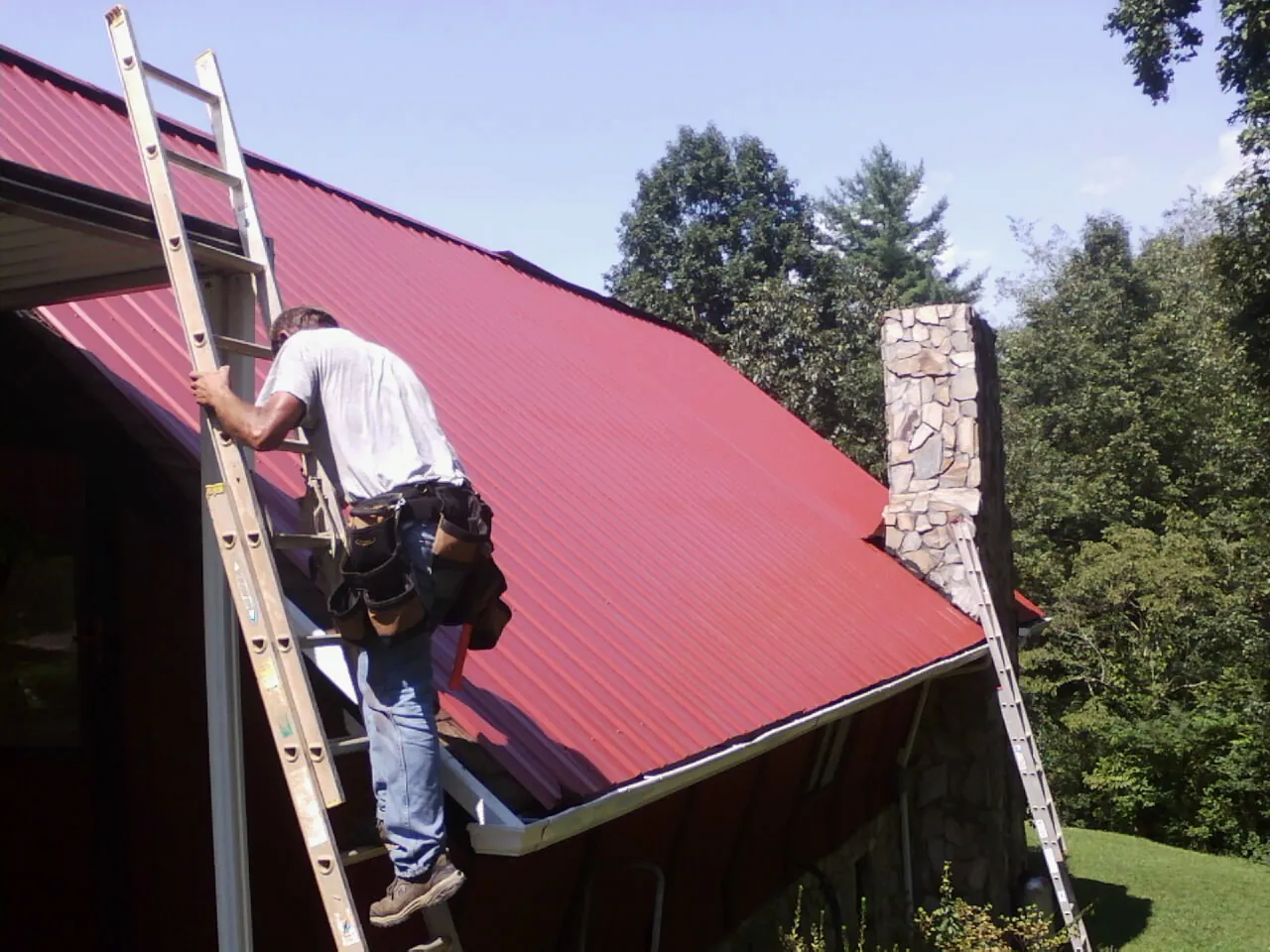 Roofer on a ladder beside a red metal roof and stone chimney — Soffit & Fascia Repair by Roofing Brothers in Haleyville